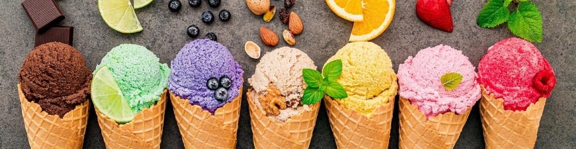 An assortment of ice cream flavors in waffle cones lined up next to one another with fruit, nuts, plants, and other indicators representing the flavor of ice cream.