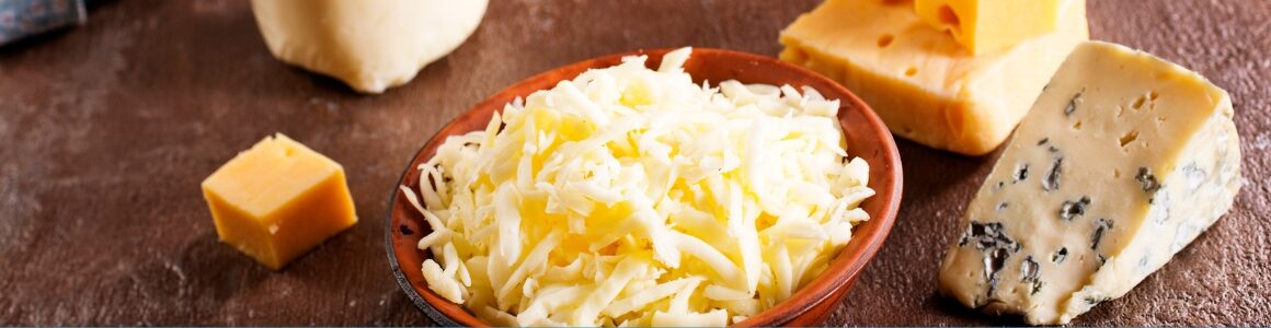 A variety of cheeses displayed on a rustic surface, including a bowl of shredded mixed milk cheese surrounded by wedges of cheddar, blue cheese, and other aged cheeses.