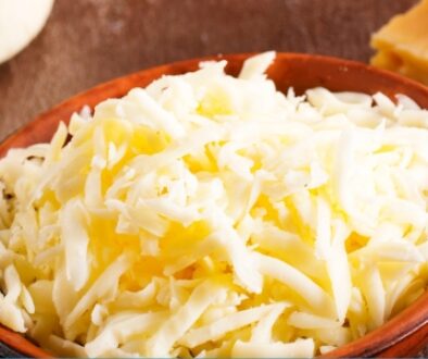 A variety of cheeses displayed on a rustic surface, including a bowl of shredded mixed milk cheese surrounded by wedges of cheddar, blue cheese, and other aged cheeses.
