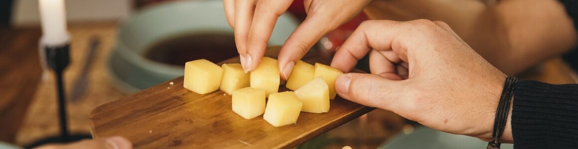Hands reaching for bite-sized cheese cubes on a wooden serving board.