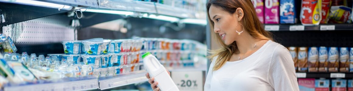 Woman examining a bottle of milk while shopping in a refrigerated dairy aisle of a grocery store.