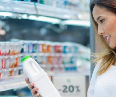 Woman examining a bottle of milk while shopping in a refrigerated dairy aisle of a grocery store.